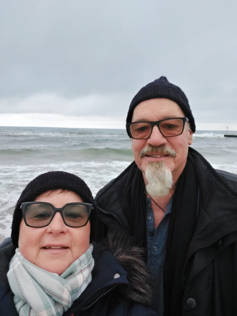Martina und Günter Görlach posieren vor einer winterlichen Ostsee in warmen Jacken und Mützen.